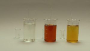A small beaker next to a larger beaker with clear, colorless liquid, and a stir rod next to another larger beaker with red clear liquid and a stir rod, and another beaker with gold-colored liquid and a stir rod