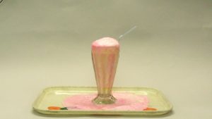 Pink-white foam spills over the edge of a soda glass on a pale tray.