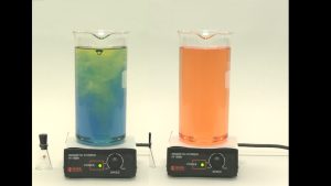 Two beakers with liquids, each on a stir plate with a black cable. The left beaker contains a mixture with a yellow-green upper layer and a blue lower layer. A stoppered Erlenmeyer flask is seen next to the hot plate. The beaker on the right contains a red-orange liquid.