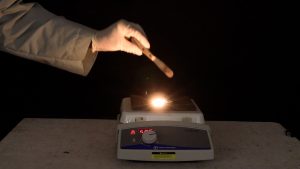 A gloved hand pours a tube with a dark solid onto a hot plate in a darkened room. An orange-white light appears on the surface of the hot plate.