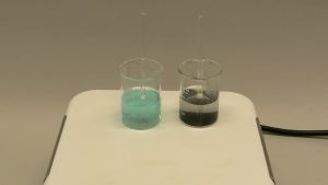 Two beakers on a pale surface: A beaker with a clear blue liquid and a beaker with a colorless liquid. A dark solid is seen at the bottom of the second beaker.