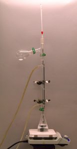 An Erlenmeyer flask connected to a glass heat exchanger and an inverted y-shaped connector with a side flask and thermometer on a white hot plate.