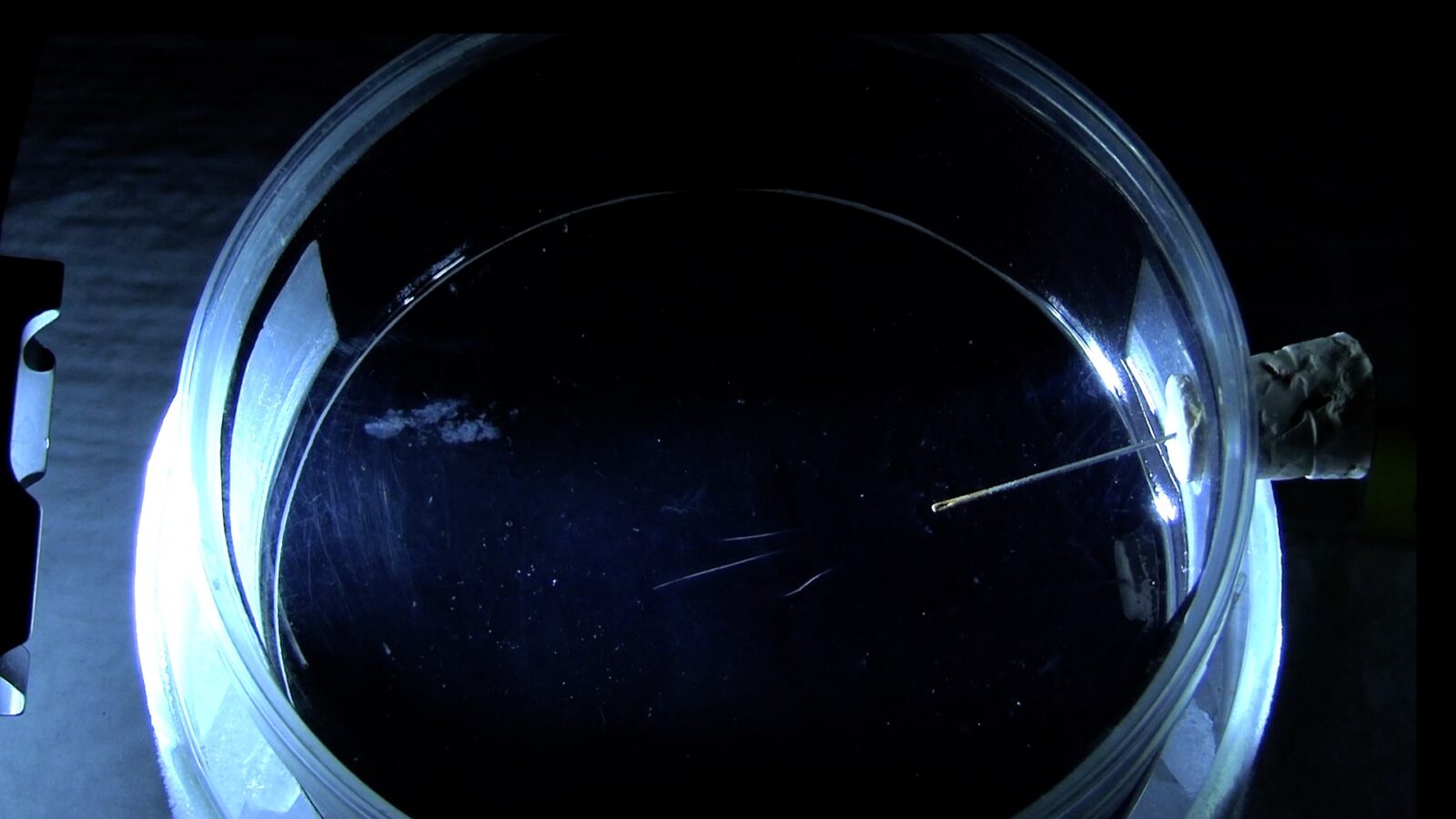 A plexiglass cylinder with an inserted needle source on a cork stopper shows white streaks of vapor denoting the presence of alpha and beta particles inside the chamber.
